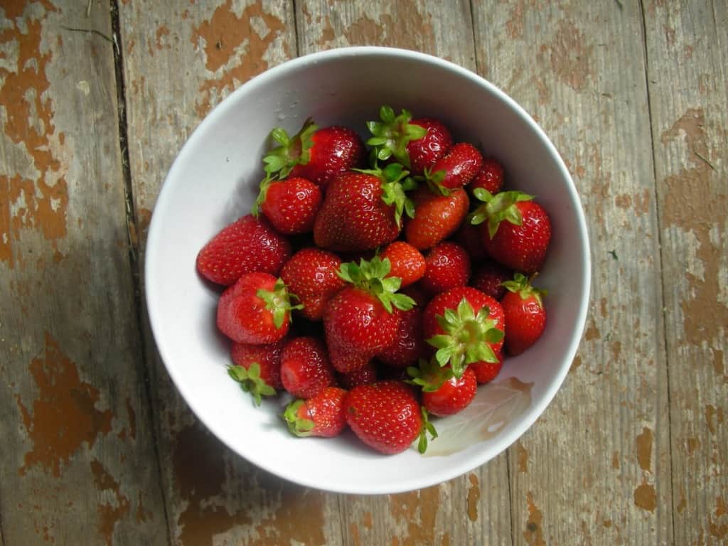 Overhead shot of a bowl of fresh strawberries in a white bowl on a wood table.