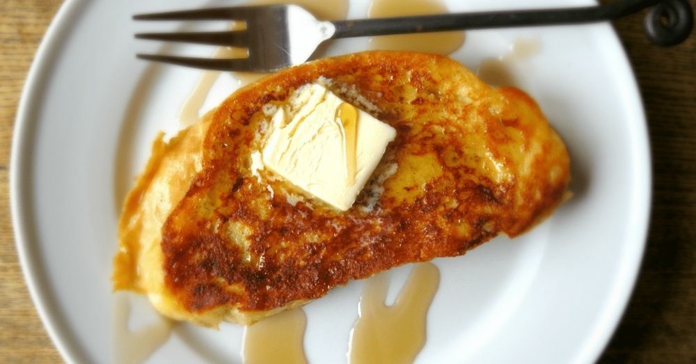Slice of french toast on a white plate with a tab of butter on top and a drizzle of maple syrup.