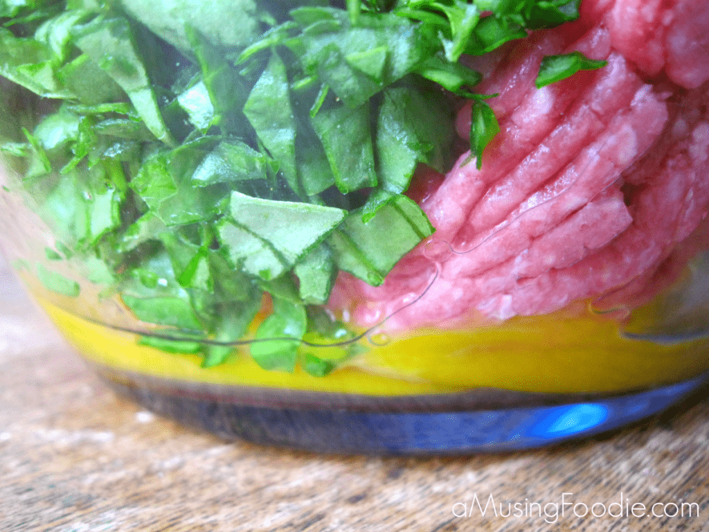 Close up shot of the side of a clear mixing bowl with egg, raw ground beef, diced spinach, sitting on top of a wooden table.