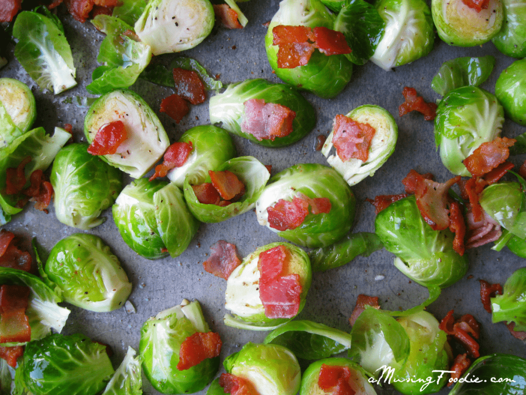 Raw brussels sprouts cut in half on a metal baking sheet, tossed with olive oil, bacon pieces, and flecks of pepper.