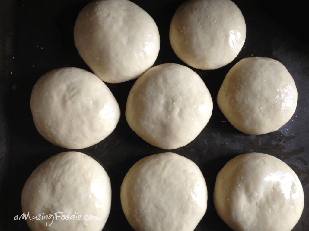Eight rounds of dough with glossy tops sitting on a dark counter.