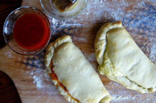 Two calzones on a wood pizza peel with tomato sauce and melted butter in glass cups to the side.