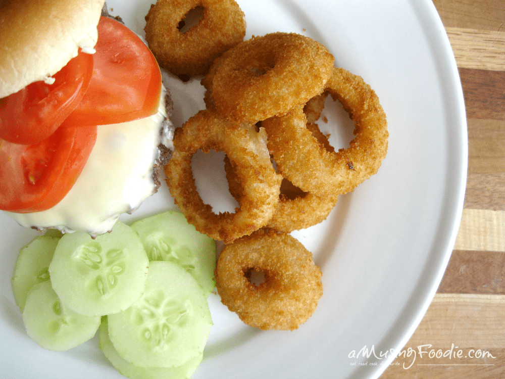 Juicy cheeseburger with crispy onion rings and fresh cucumber slices on a white plate.