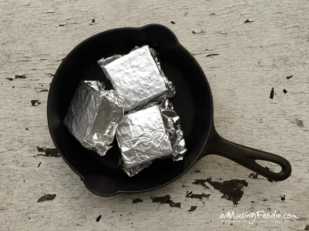 S'mores wrapped tightly in foil packets, sitting in a black cast iron skillet on top of a white table with peeling paint.