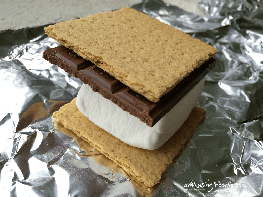 Chocolate bar and marshmallow sandwiched between two graham cracker cookies on top of aluminum foil.
