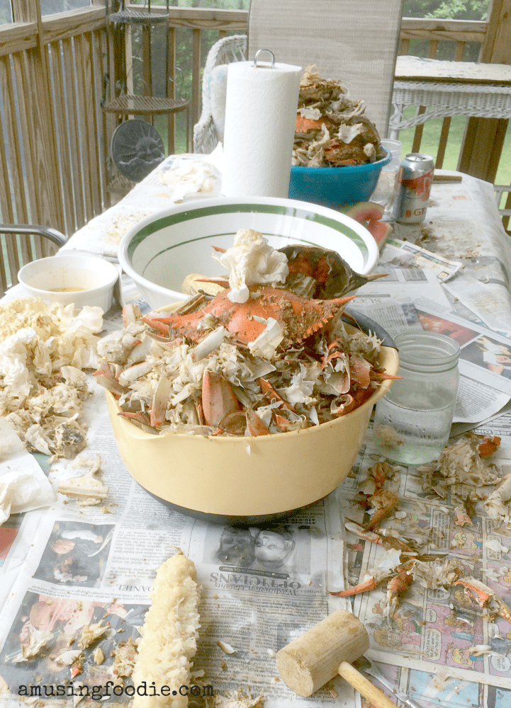 Table in a screen porch covered with newspapers, empty blue crab shells, eaten corn on the cob and paper towels.