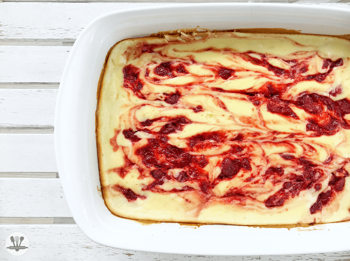 Overhead shot of a white baking dish with strawberry swirls in a cooked cheesecake, on a white table.