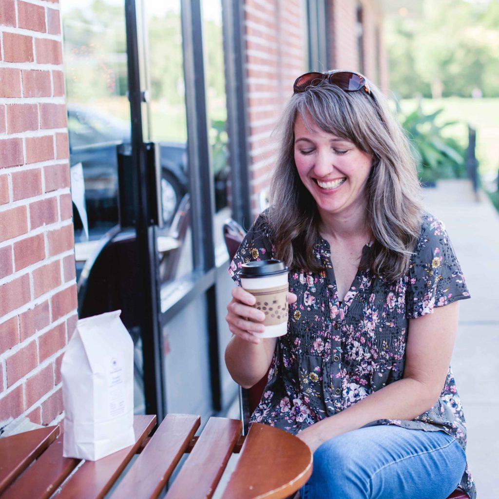 Woman laughing while holding a coffee at an outside table.
